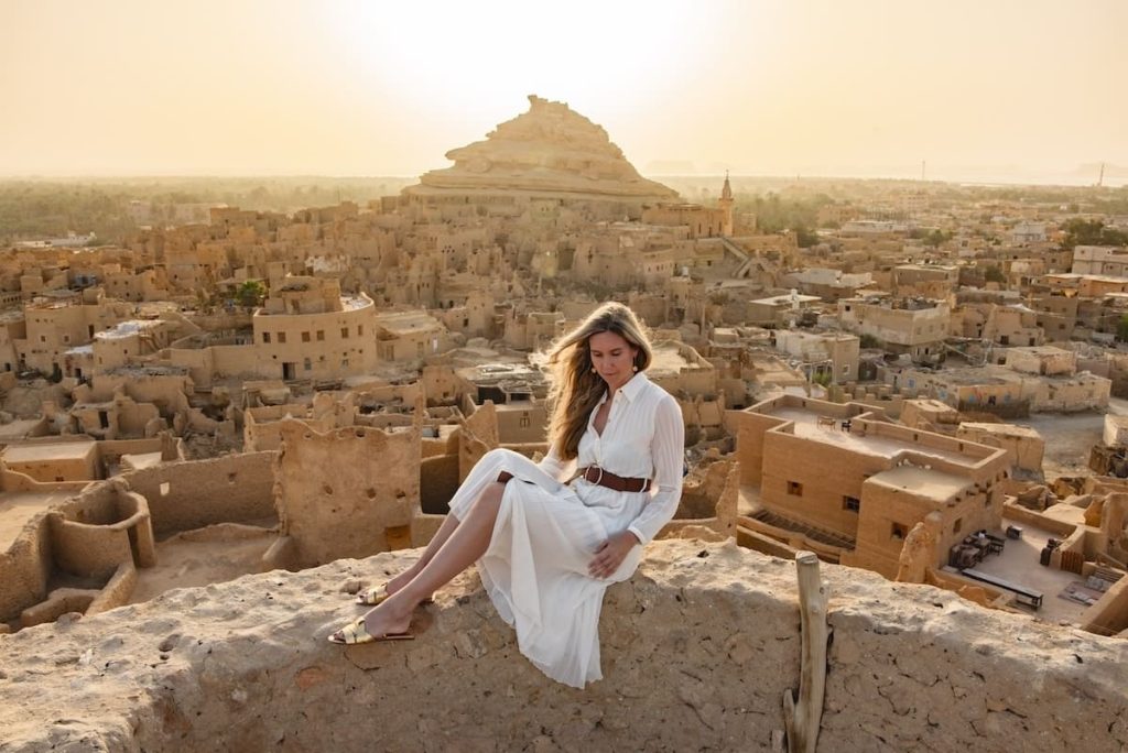 A woman in a white dress sitting on a wall, overlooking the ancient mud-brick buildings of Siwa Oasis at sunset.