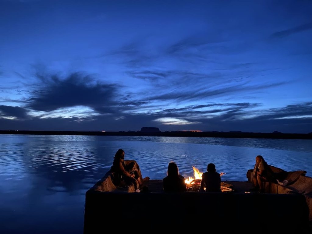 People gathered around a campfire by a serene lake at twilight, with a dramatic sky and distant hills in the background.