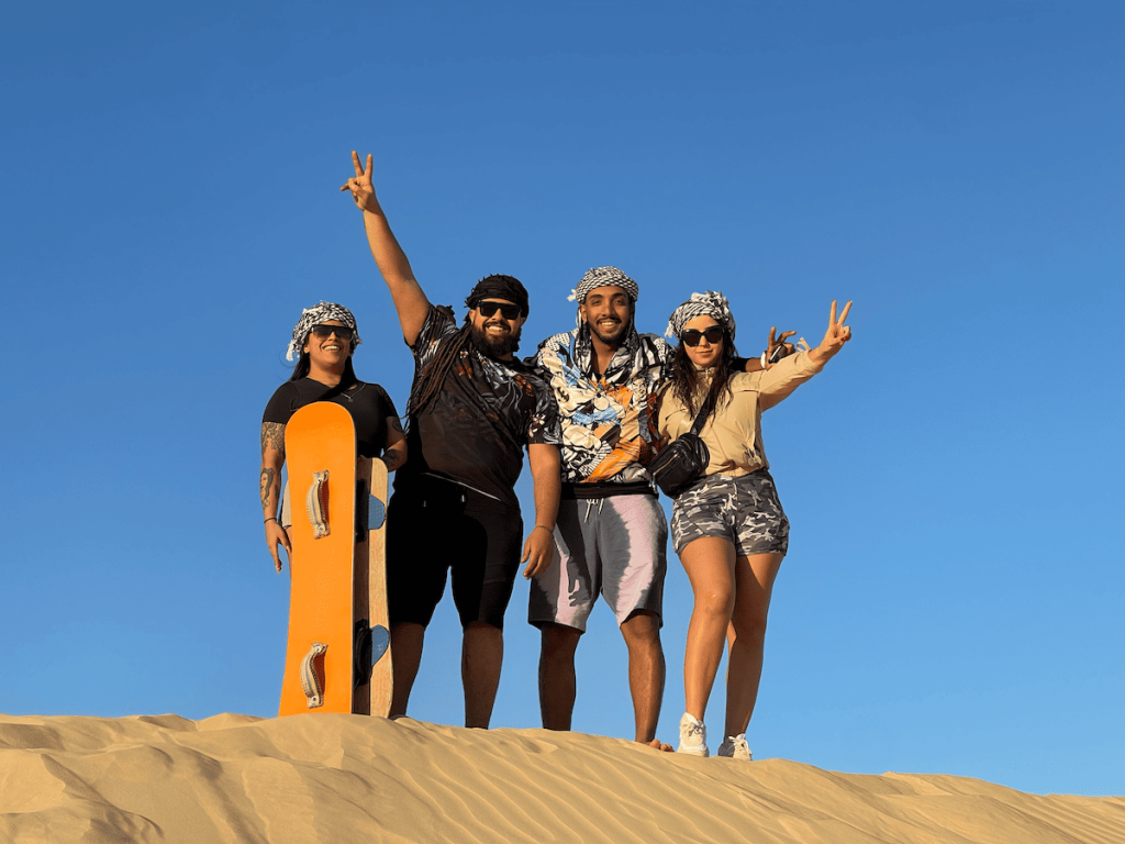 Four friends posing with a sandboard, celebrating on a vast sand dune under a clear blue sky.