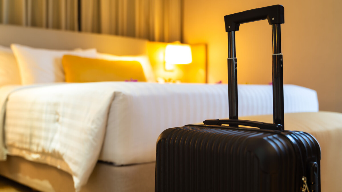 "Close-up of a black suitcase with an unfocused background of a hotel room with a well-made bed.
