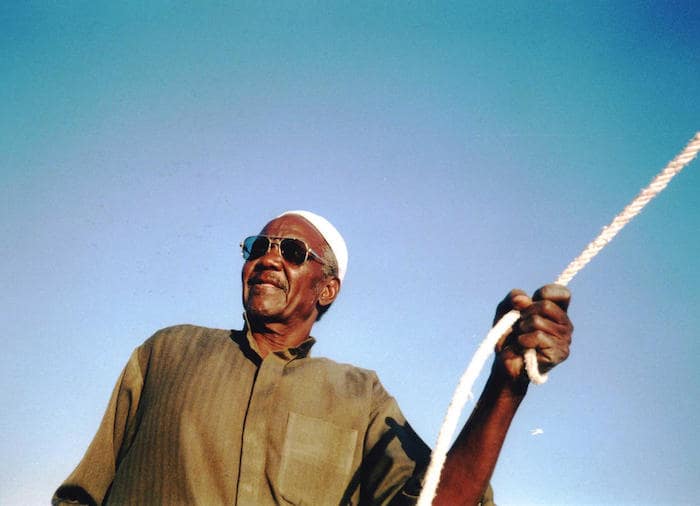 A Nubian felucca captain in traditional attire, holding a rope with a clear blue sky in the background.