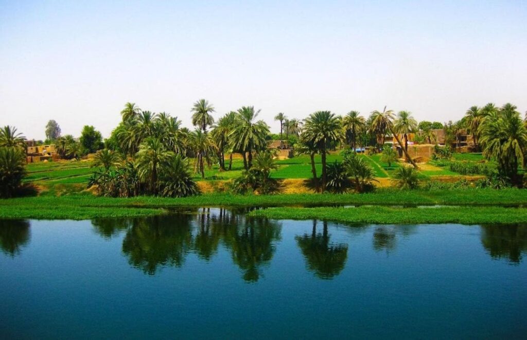 Lush greenery and palm trees reflect on the calm waters of the Nile River.