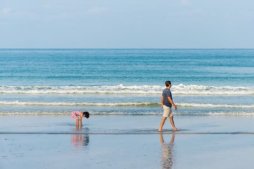 A man walking along the beach with a child bending down to the sand near the water's edge.