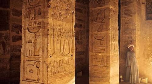 Interior of Amada Temple in Aswan showing detailed hieroglyphs on columns with a local guardian standing nearby.