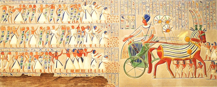 Colorful ancient Egyptian wall relief depicting a pharaoh riding a chariot, followed by rows of hieroglyphs and figures.