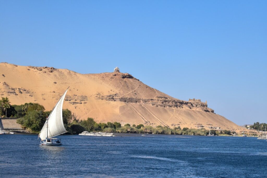 Traditional Egyptian sailboat, known as a felucca, on the Nile River with sandy hills in the background.