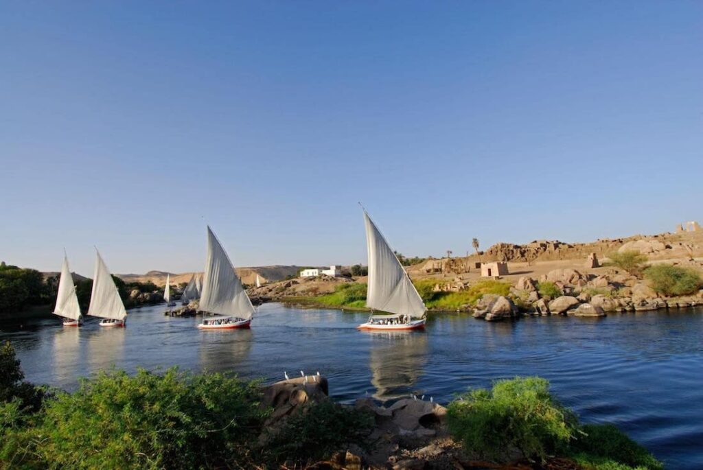 Traditional feluccas sailing on the Nile River with a backdrop of rocky outcrops and greenery.