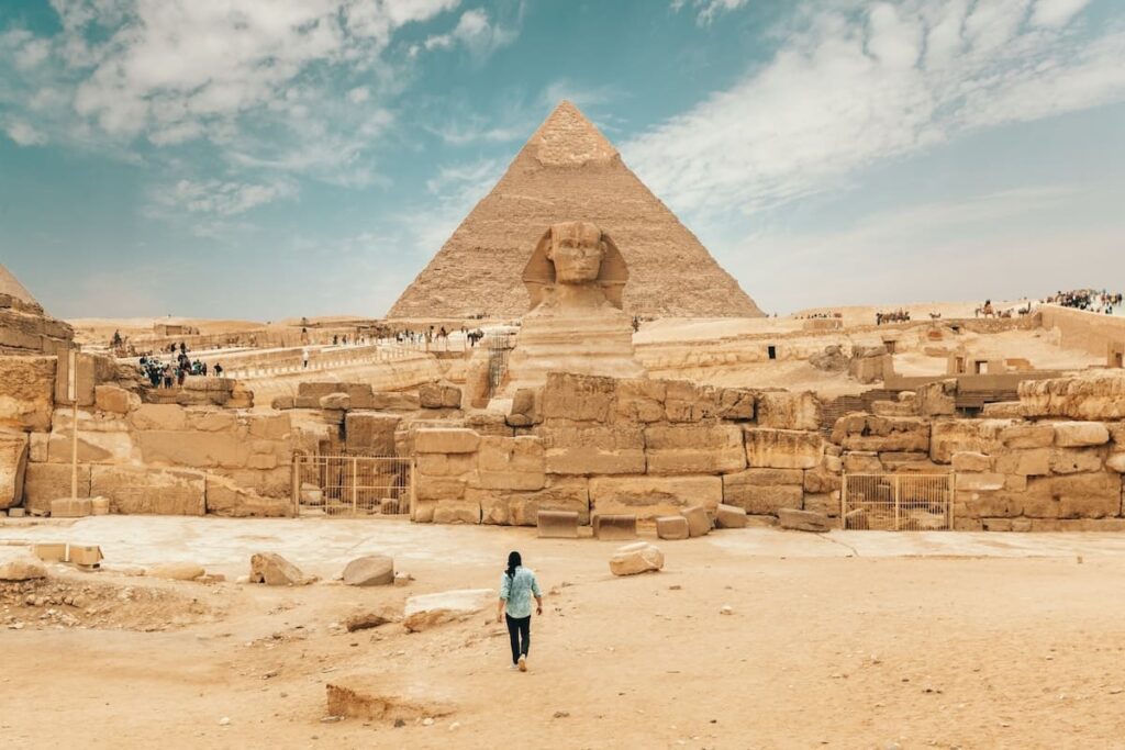 A person walking towards the Great Sphinx with the Pyramid of Khafre in the background.