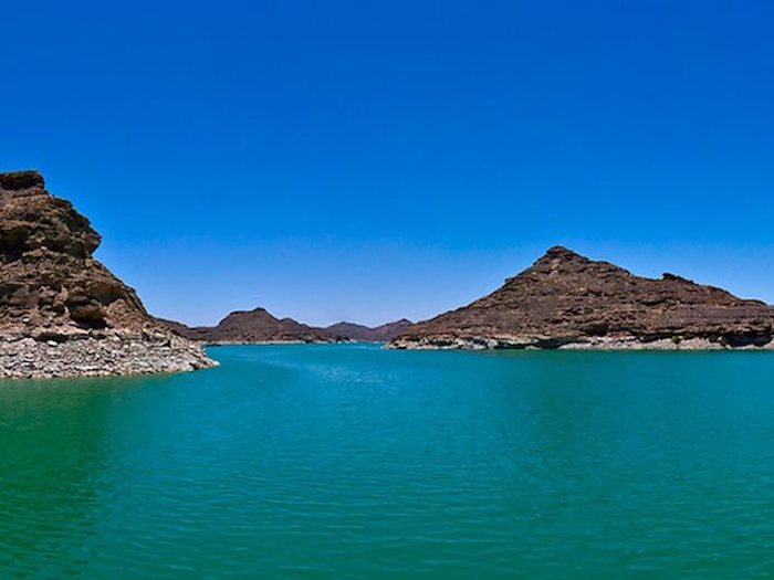 A tranquil view of the turquoise waters of Lake Nasser with surrounding barren rocky hills under a clear blue sky.