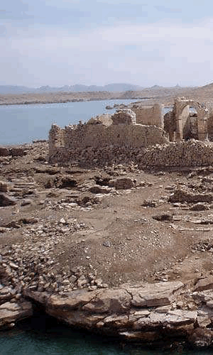 The ancient stone ruins of Qasr Ibrim overlooking a lake in Aswan, Egypt, under a hazy sky.
