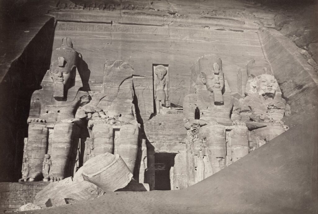 Black and white photograph of the Great Temple of Abu Simbel, featuring four giant seated statues of Pharaoh Ramesses II, partially covered in sand.