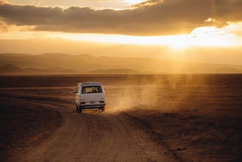 A white van traveling down a dusty road at sunset in a vast, open landscape.