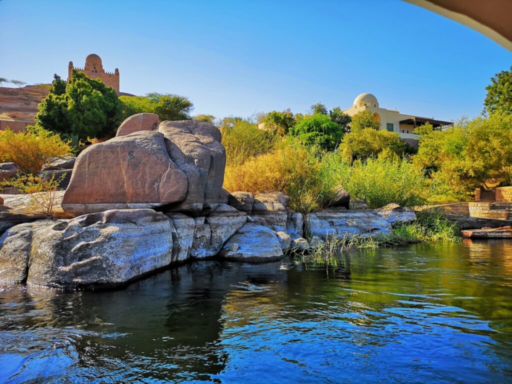 Tranquil waters of the Nile River flowing by rocks and greenery with a dome structure in the distance.
