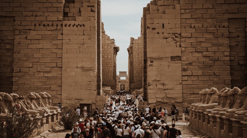 "Crowds of tourists walking through the ancient Karnak Temple complex with rows of ram-headed sphinx statues lining the path."