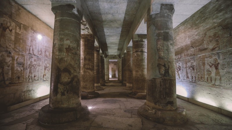 "Interior view of an ancient Egyptian temple corridor with columns and hieroglyphic carvings illuminated by spotlights."