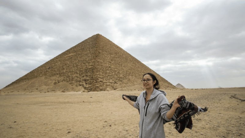 "A person stands in front of the Great Pyramid of Giza on a cloudy day, holding a jacket in one hand."