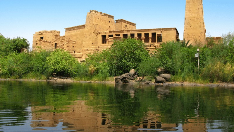 "The Temple of Philae reflected in the Nile River, surrounded by lush greenery under a clear blue sky."