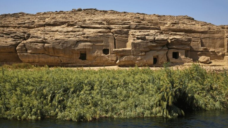 - "Rock-cut tombs by the Nile riverbank surrounded by lush greenery."