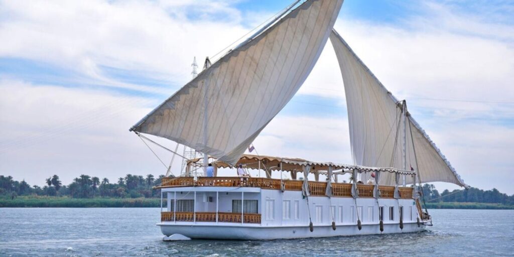 "A traditional Dahabiya sailboat with large white sails on the Nile River under a cloudy sky."