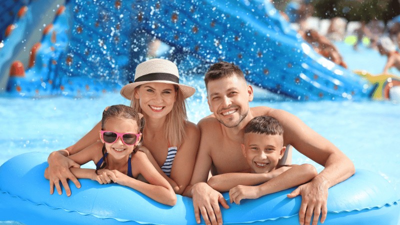 A family of four smiling and hugging on a blue inflatable raft at a water park with water splashes in the background
