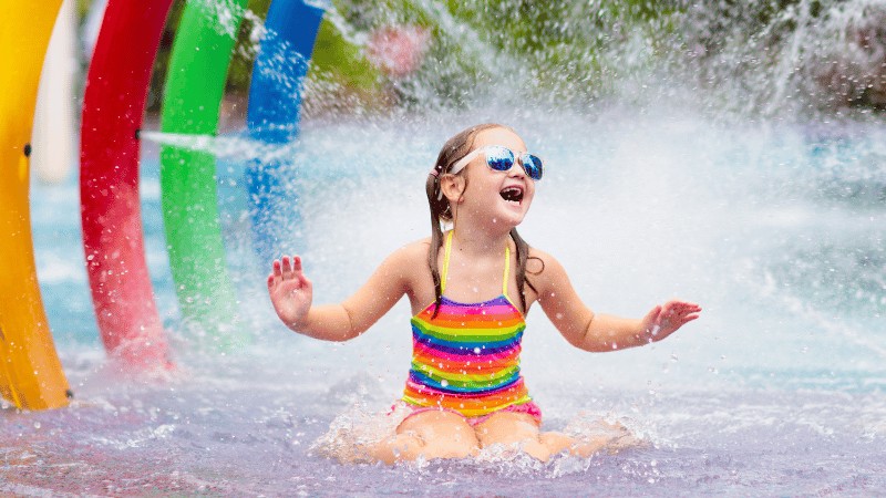 A young girl with sunglasses laughing joyfully as she slides down a colorful water slide into a splash pool."A young girl in a rainbow-striped swimsuit enjoys a water slide at an aqua park, expressing sheer delight surrounded by vibrant colors and splashes of water