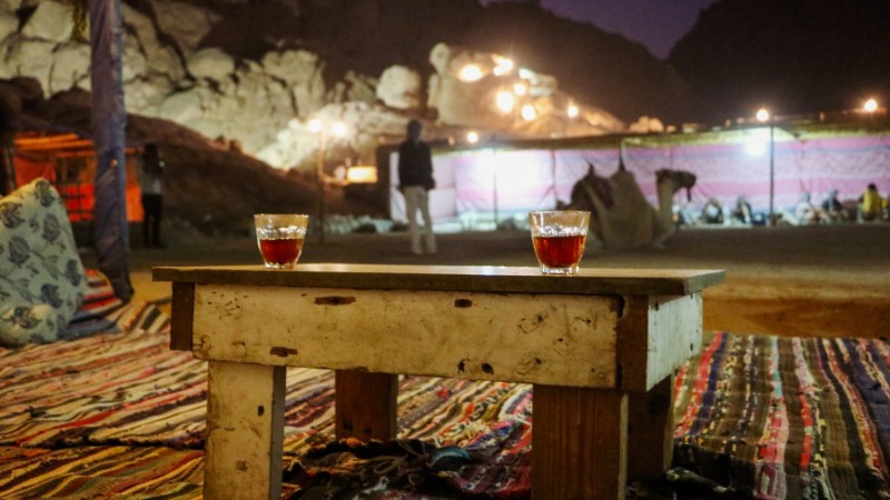 "Two glasses of tea on a wooden table in a Bedouin camp at dusk, with dim lights and a camel in the background."
