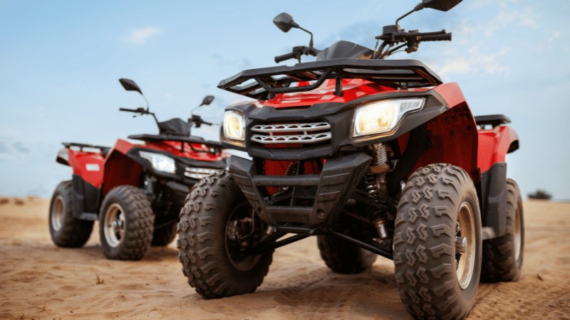 "Two red all-terrain vehicles parked on sandy terrain under a clear sky."