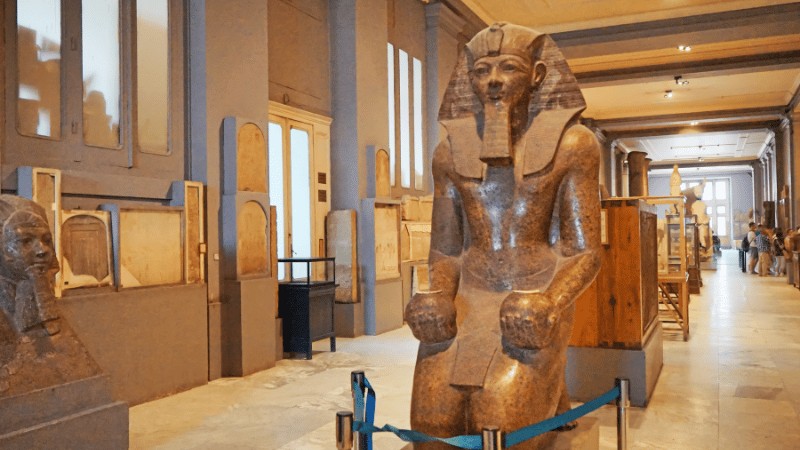 "A large, seated statue of a pharaoh with a headdress in the foreground of an exhibit hall lined with Egyptian artifacts."