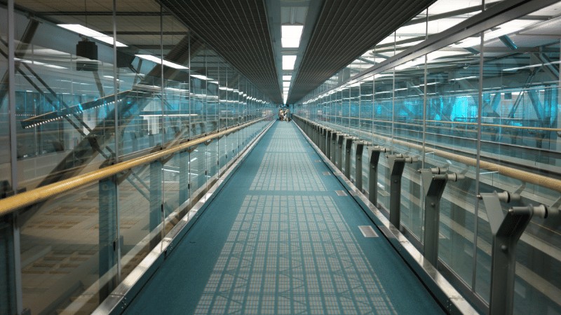 "Interior view of a modern airport walkway with glass walls and blue-tinted windows."