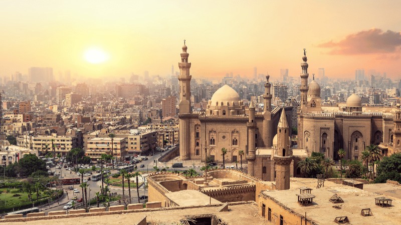 "A panoramic view of Islamic Cairo with historic mosques and minarets against a sunset sky."