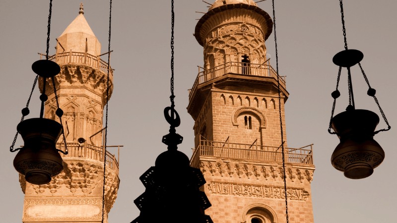 "Silhouette of intricately designed Islamic minarets with hanging traditional lanterns against a clear sky."