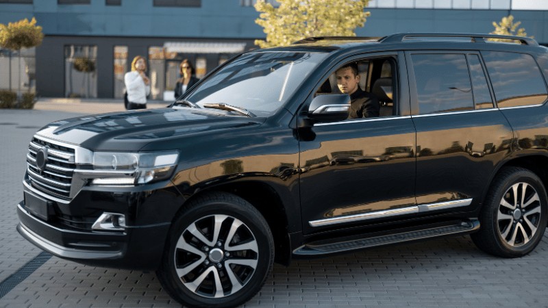"A shiny black luxury SUV parked on a city street, with a driver at the wheel and pedestrians in the background."