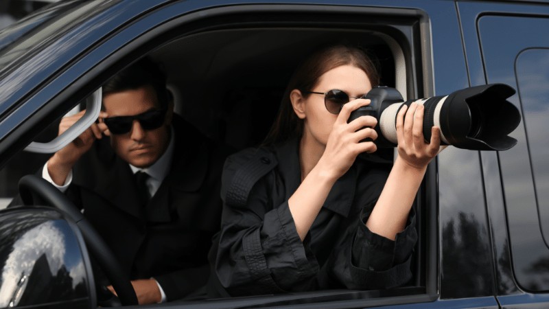 Two people in a car, one using a camera and the other watching out, both wearing sunglasses.