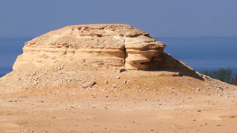 "A lone eroded sandstone formation stands against a clear sky, overlooking a tranquil sea in the background."