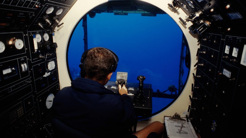 A pilot operating a submersible's controls while submerged in the ocean, viewed from inside the cockpit