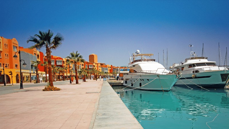 "Luxury yachts moored at a marina with orange-hued buildings and palm trees under a clear blue sky."