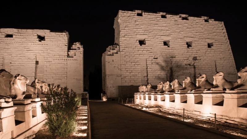 "A night view of the entrance to Karnak Temple, lined with Sphinx statues, illuminated by artificial lighting."