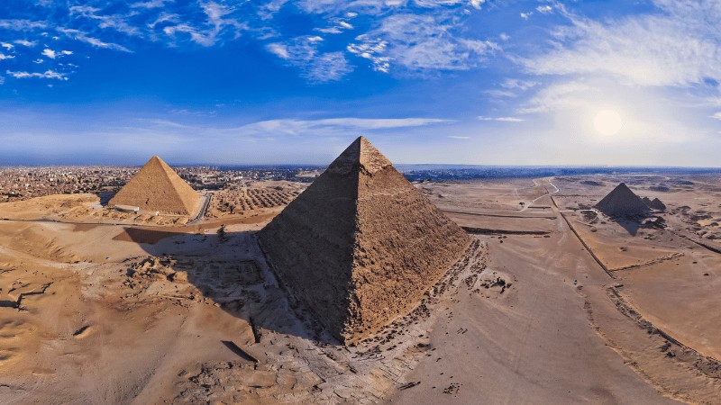 "A panoramic view of the Giza Pyramid Complex against a clear blue sky with the sun shining brightly."