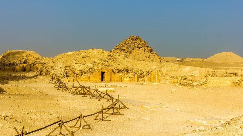"Ruins of small step pyramids and ancient tombs with remnants of walls in the Egyptian desert under a clear blue sky."