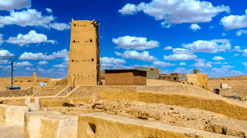 "Ancient clay brick watchtower rising above a desert town under a blue sky with clouds."