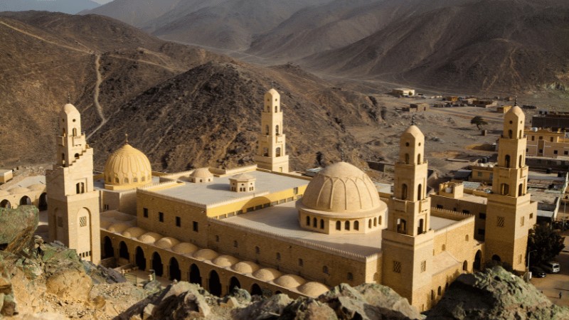 "A majestic mosque with multiple minarets and a golden dome nestled in a desert valley surrounded by mountains."