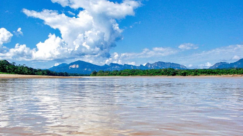 A wide river with muddy waters flows in the foreground, flanked by lush greenery, with a backdrop of distant mountains under a blue sky dotted with clouds.