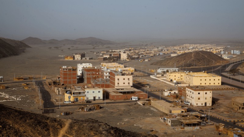 "A view of a developing town on the edge of a desert with buildings in the foreground and expansive sandy terrain in the background."