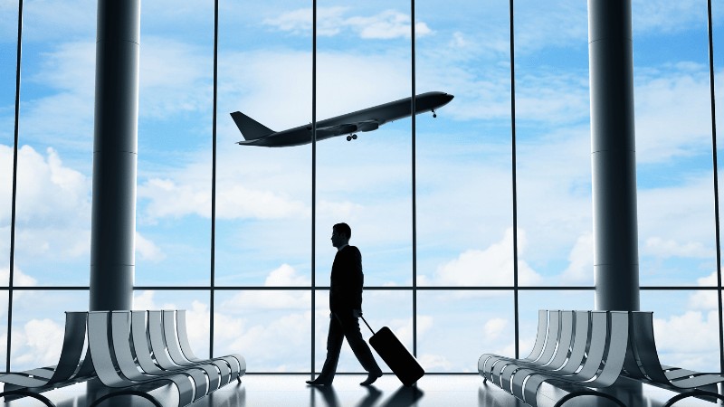 "Silhouette of a person walking in an airport terminal with a suitcase, a flying airplane visible through the large windows."