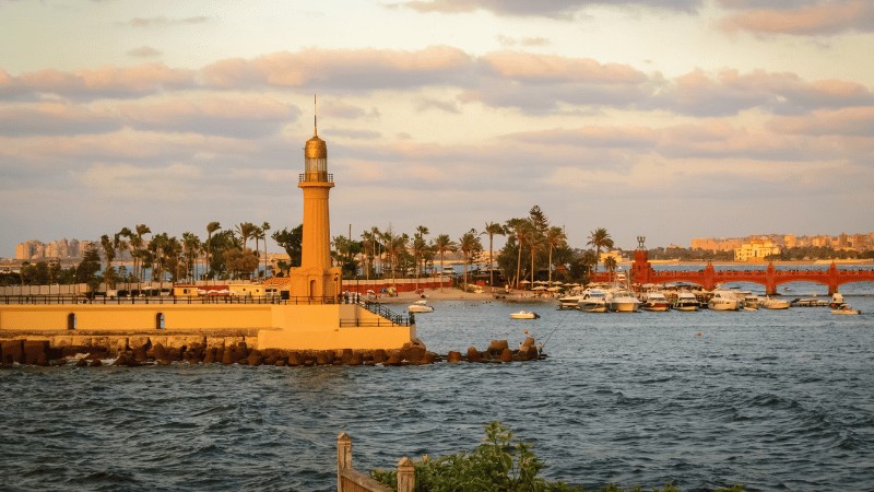 "A lighthouse stands at the edge of a harbor with boats and palm trees during sunset."