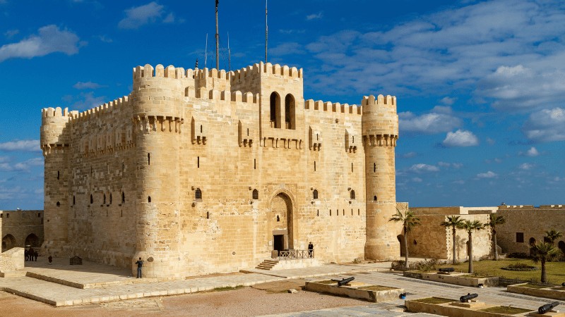 e Citadel of Qaitbay, a formidable medieval fortress with stone walls and watchtowers, stands under a clear blue sky in Alexandria, Egypt.