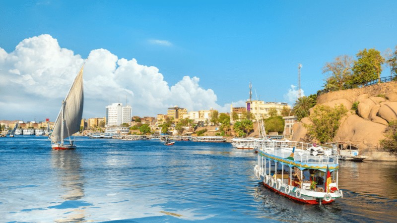 "Traditional felucca sailing boat and a tourist riverboat on the blue waters of the Nile River with the Aswan city skyline in the background under a sunny sky with puffy clouds."