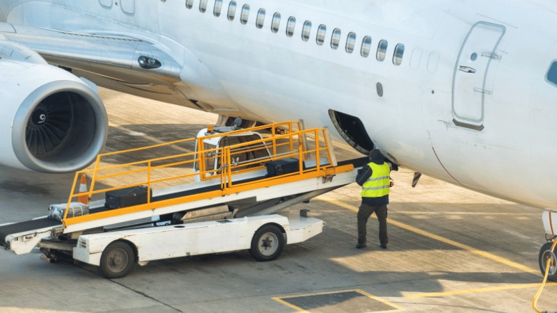 "An airport ground crew member loading luggage onto a commercial airplane via a conveyor belt vehicle."