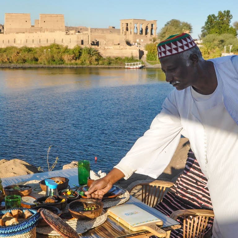 Man setting a traditional table by the Nile River with Philae Temple in the background.