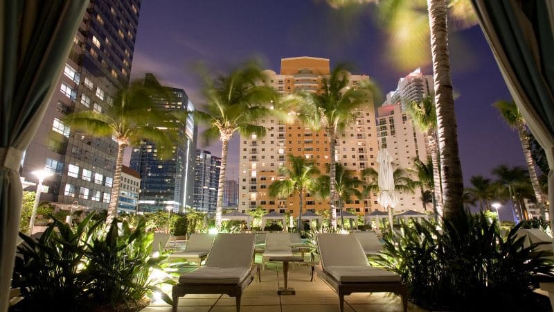 Poolside loungers in an urban hotel at night, surrounded by illuminated high-rises and palm trees.